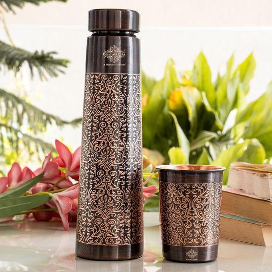 Decorative water bottle and cup with intricate patterns on a table with flowers and books in the background.