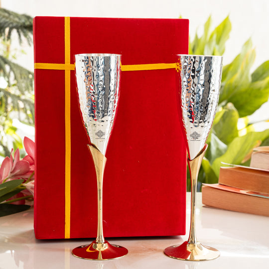 Two silver champagne flutes with gold bases in front of a red gift box on a white surface with greenery in the background.