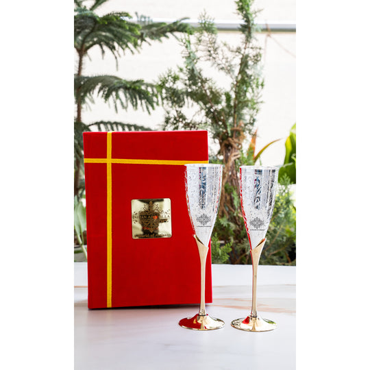 Red gift box with gold ribbon next to two champagne flutes on a white surface with greenery in the background.